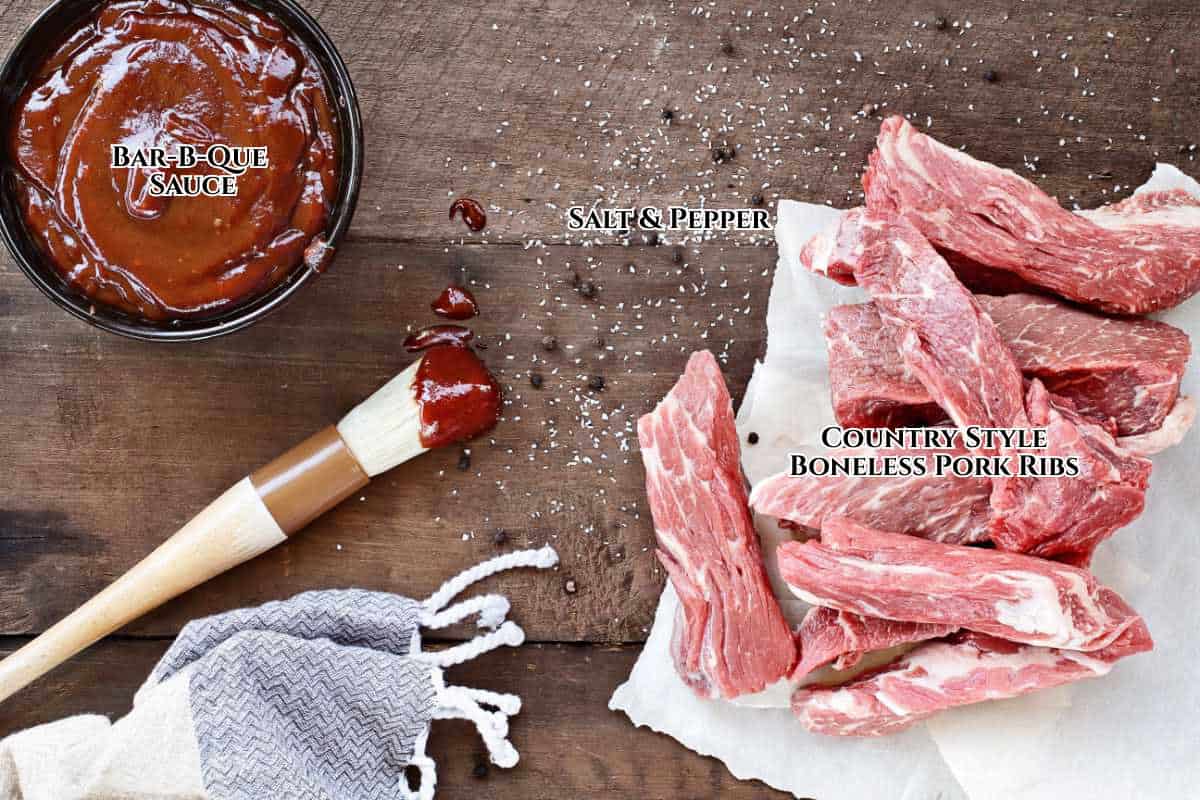 Country style boneless pork ribs on parchment paper with salt and pepper sprinkled nearby, a bowl of barbecue sauce, and a basting brush sit ready on a rustic wooden surface.