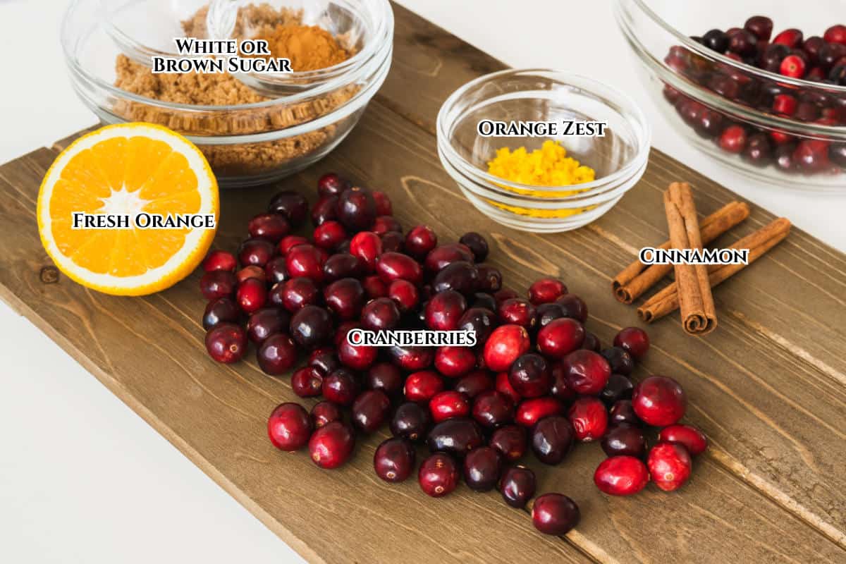A wooden board displays ingredients for homemade cranberry orange sauce: fresh cranberries, an orange half, cinnamon sticks, bowls of brown sugar, orange zest, and cranberries, each labeled with text.