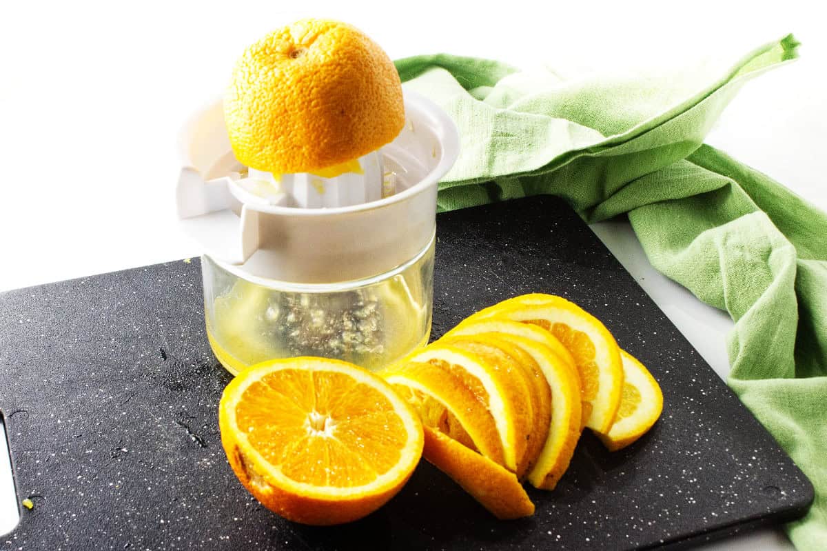 A manual citrus juicer with half an orange on top, a jar collecting juice below, and several orange slices beside it on a black cutting board—perfect prep for homemade cranberry orange sauce. A green cloth is in the background.