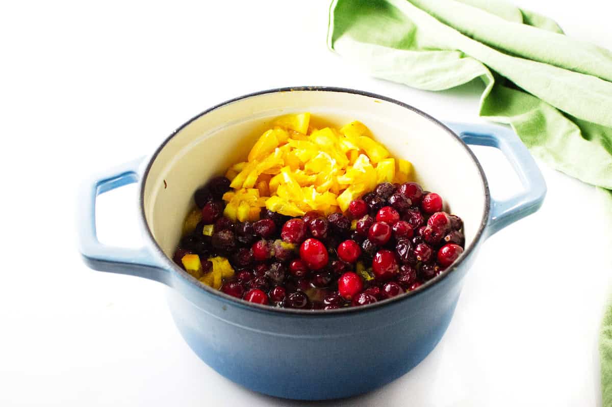 A blue pot filled with fresh cranberries, chopped yellow bell peppers, and zesty homemade cranberry orange sauce sits on a white surface with a green cloth nearby.