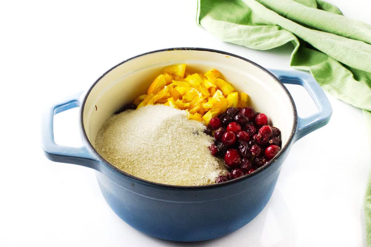 A blue pot holds chopped yellow fruit, whole cranberries, and a mound of granulated sugar—ingredients for homemade cranberry orange sauce—arranged separately. A green cloth rests nearby on a white surface.