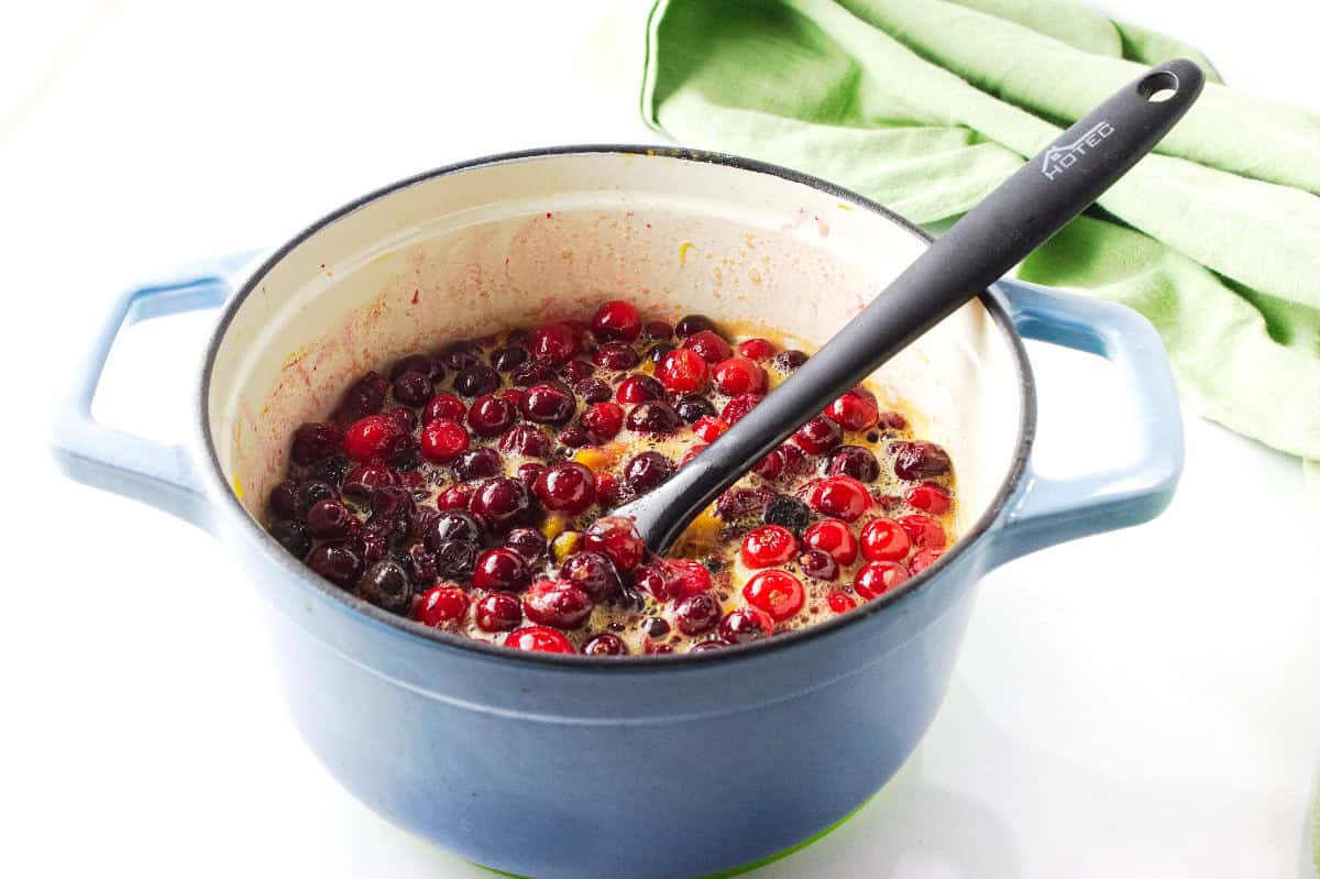 A blue pot filled with cranberries being cooked for a homemade cranberry orange sauce, with a black spoon resting inside. A green cloth is in the background on a white surface.