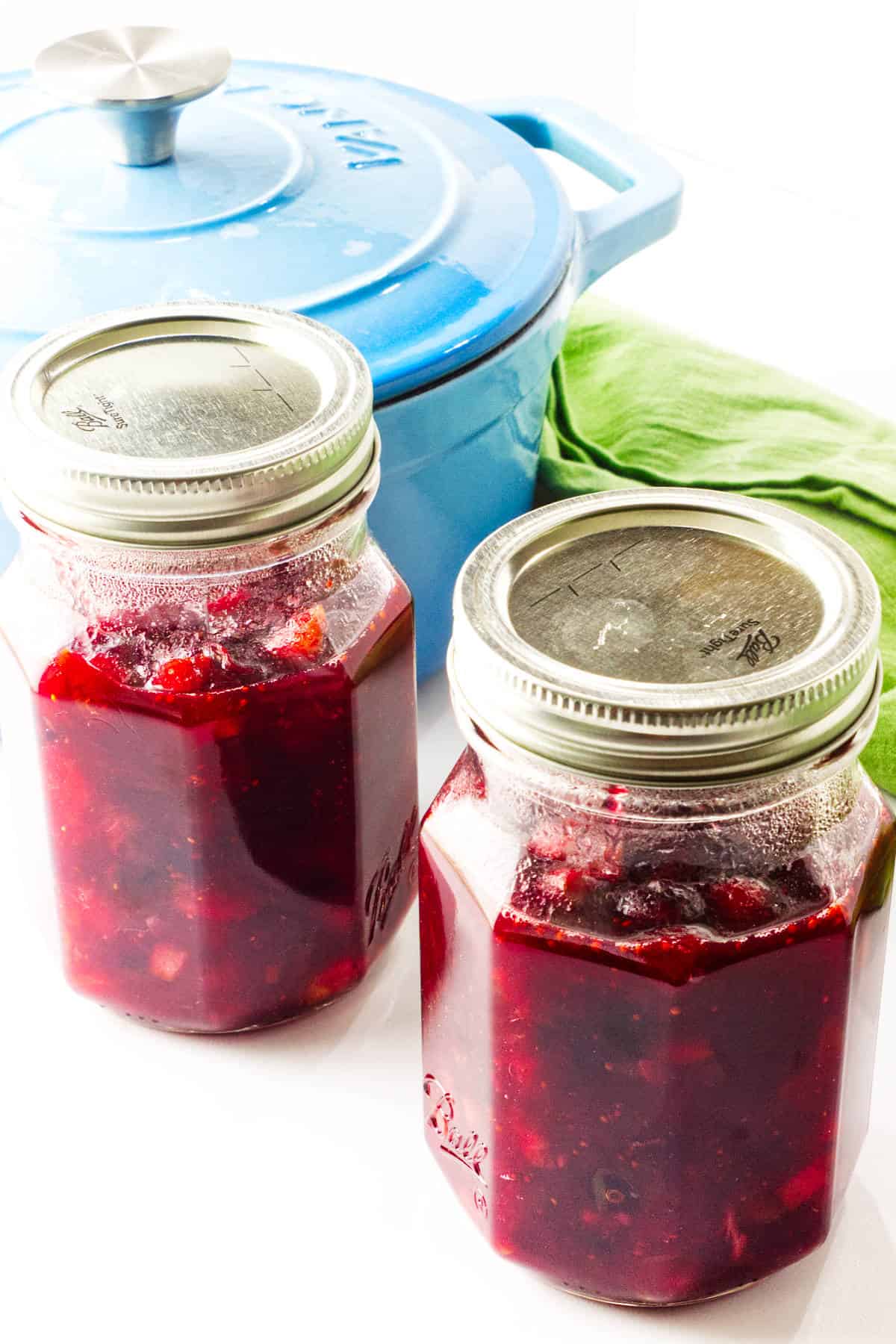 Two glass jars filled with homemade cranberry orange sauce are placed in front of a blue Dutch oven and a green kitchen towel on a white surface.