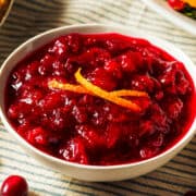 A white bowl filled with chunky homemade cranberry orange sauce, garnished with thin strips of orange zest, sits on a striped cloth.