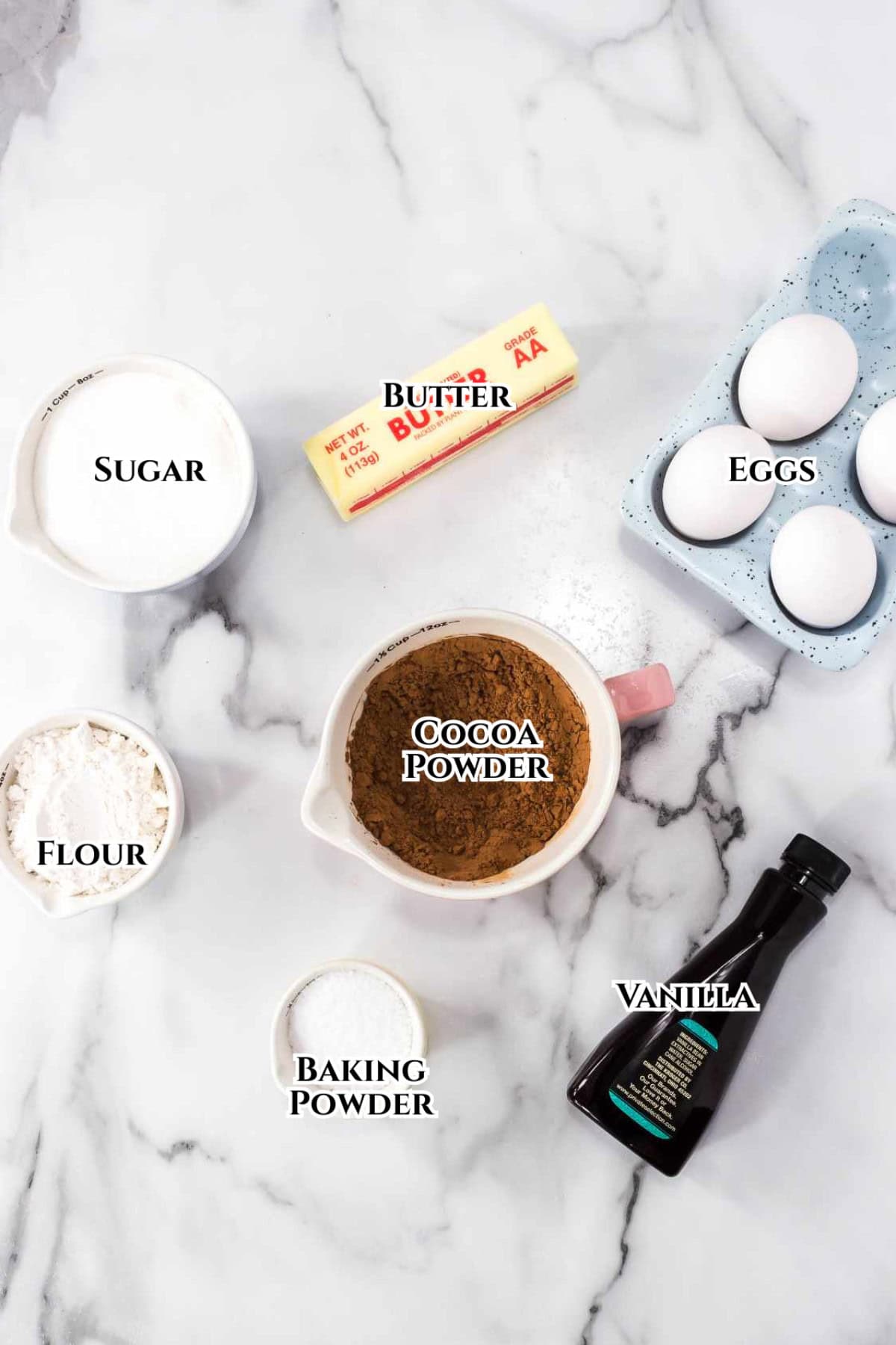 Baking ingredients on a marble surface for one bowl brownies: a stick of butter, three eggs in a carton, a bottle of vanilla, a bowl of cocoa powder, sugar, flour, and baking powder, each labeled with text.