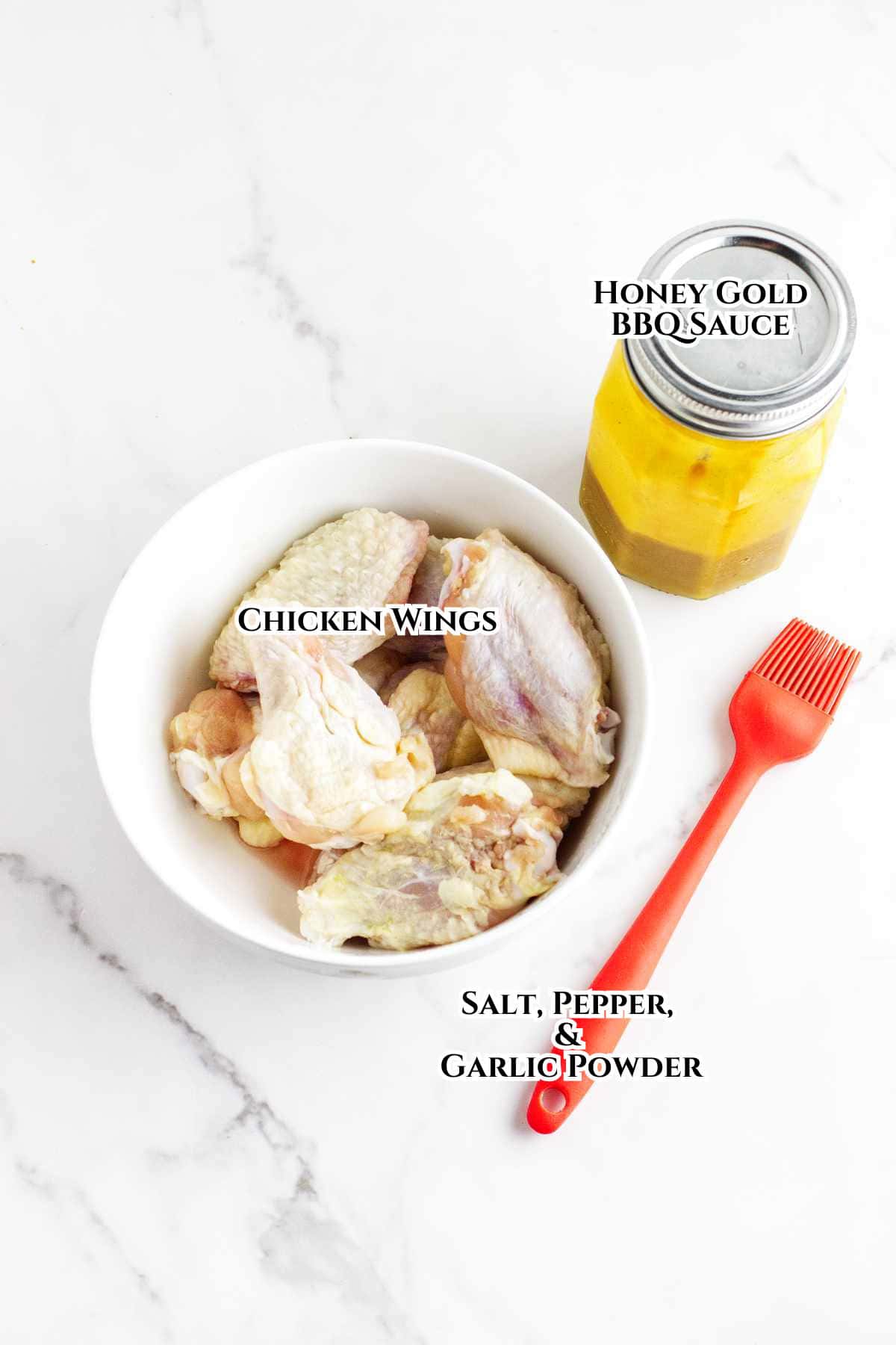 A bowl of raw chicken wings, a jar of honey gold BBQ sauce, and a small pile of salt, pepper, and garlic powder with a red basting brush on a white marble surface.