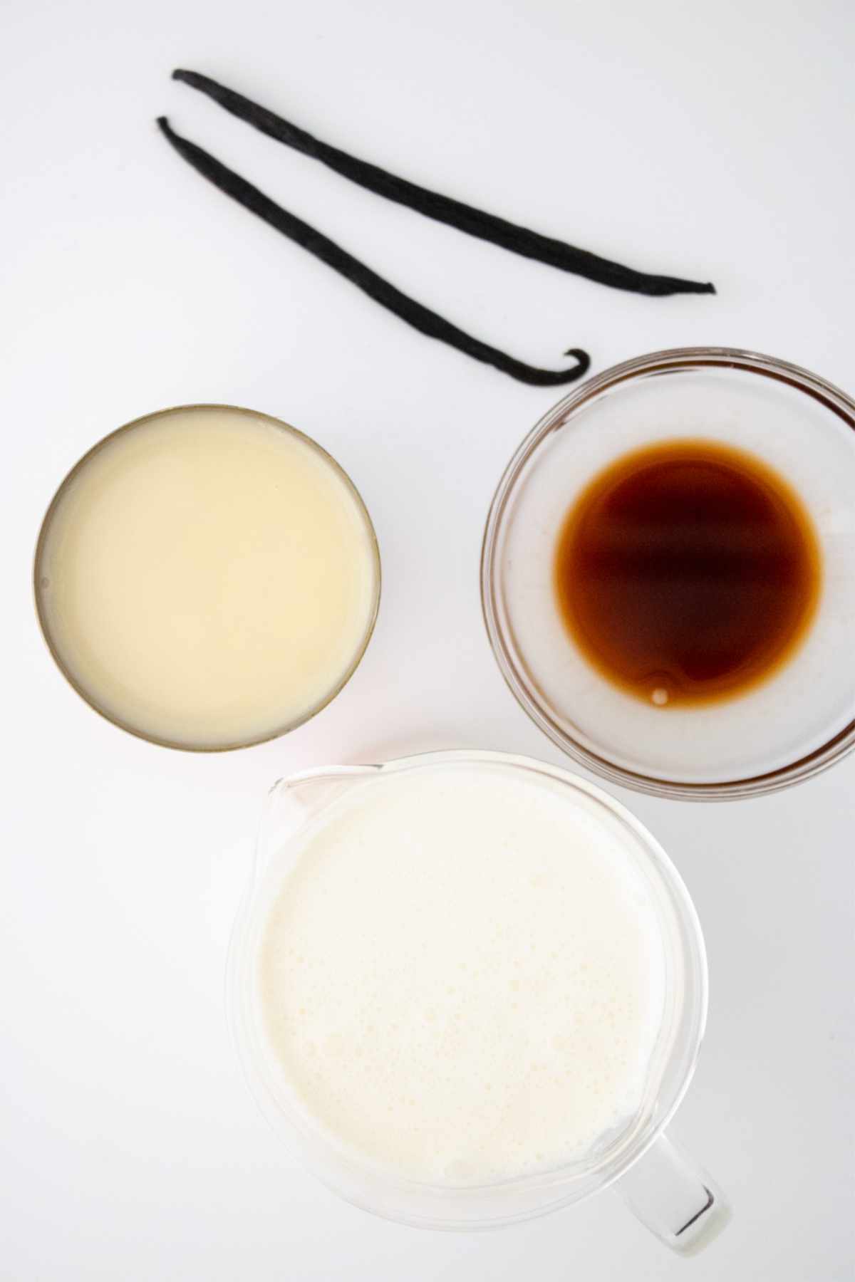 Three ingredients on a white surface: two vanilla beans, a glass of sweetened condensed milk, a glass cup of coffee or espresso, and a glass pitcher—perfect for making rich ice cream maker vanilla ice cream at home.