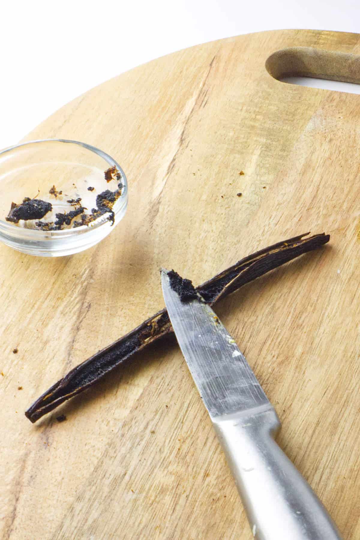 A knife scraping seeds from a split vanilla bean on a wooden cutting board, with a small glass bowl of vanilla seeds nearby—perfect for adding pure flavor to your next ice cream maker vanilla ice cream creation.
