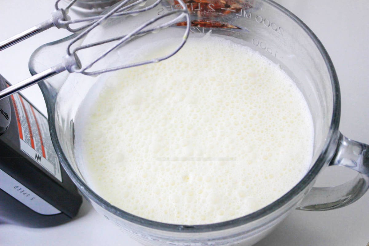 A glass mixing bowl filled with a frothy, whipped cream mixture rests beside an ice cream maker, ready to be turned into homemade vanilla ice cream. An electric hand mixer with beaters sits on the bowl’s edge.