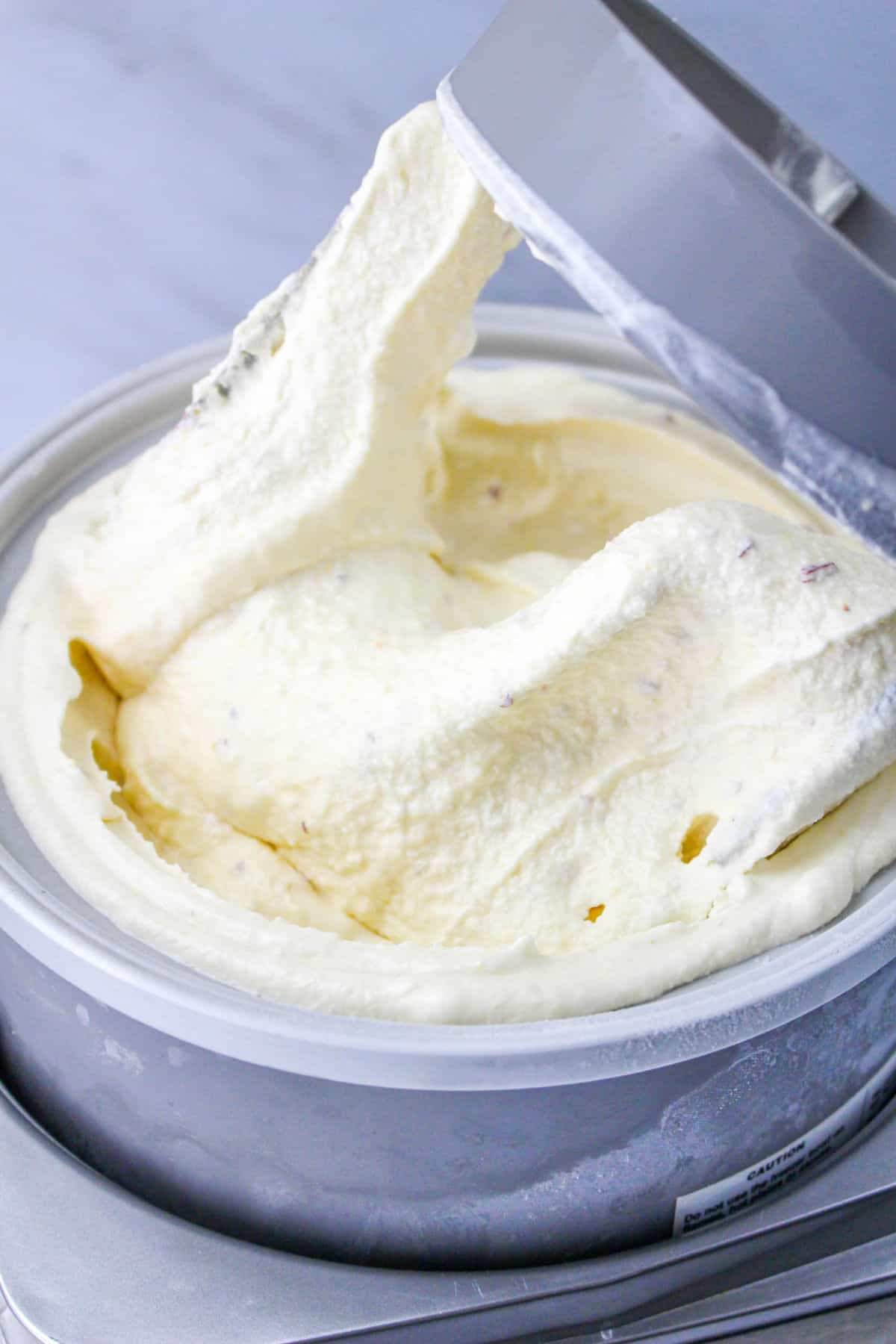 A close-up of creamy vanilla gelato being scooped from a metal container, with the soft, smooth texture visible and the lid partially open.