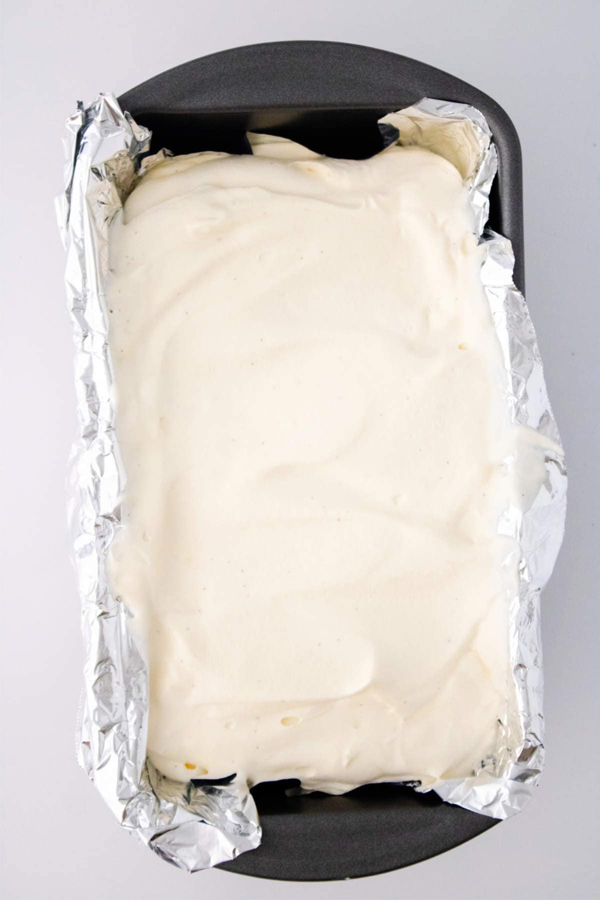 A loaf pan lined with aluminum foil, filled with smooth, creamy white cheesecake batter—so luscious it looks like it could have been made in an ice cream maker for vanilla ice cream—ready to be baked.