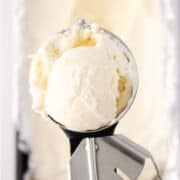 A close-up of a metal ice cream scoop holding a round scoop of vanilla ice cream, freshly made in an ice cream maker.