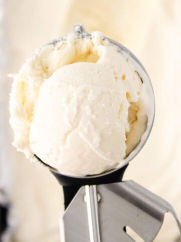 A close-up of a metal ice cream scoop holding a round scoop of vanilla ice cream, freshly made in an ice cream maker.