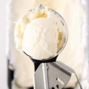 A metal ice cream scoop holds a rounded scoop of creamy vanilla ice cream, freshly made with an ice cream maker, with more vanilla ice cream visible in a container in the blurred background.