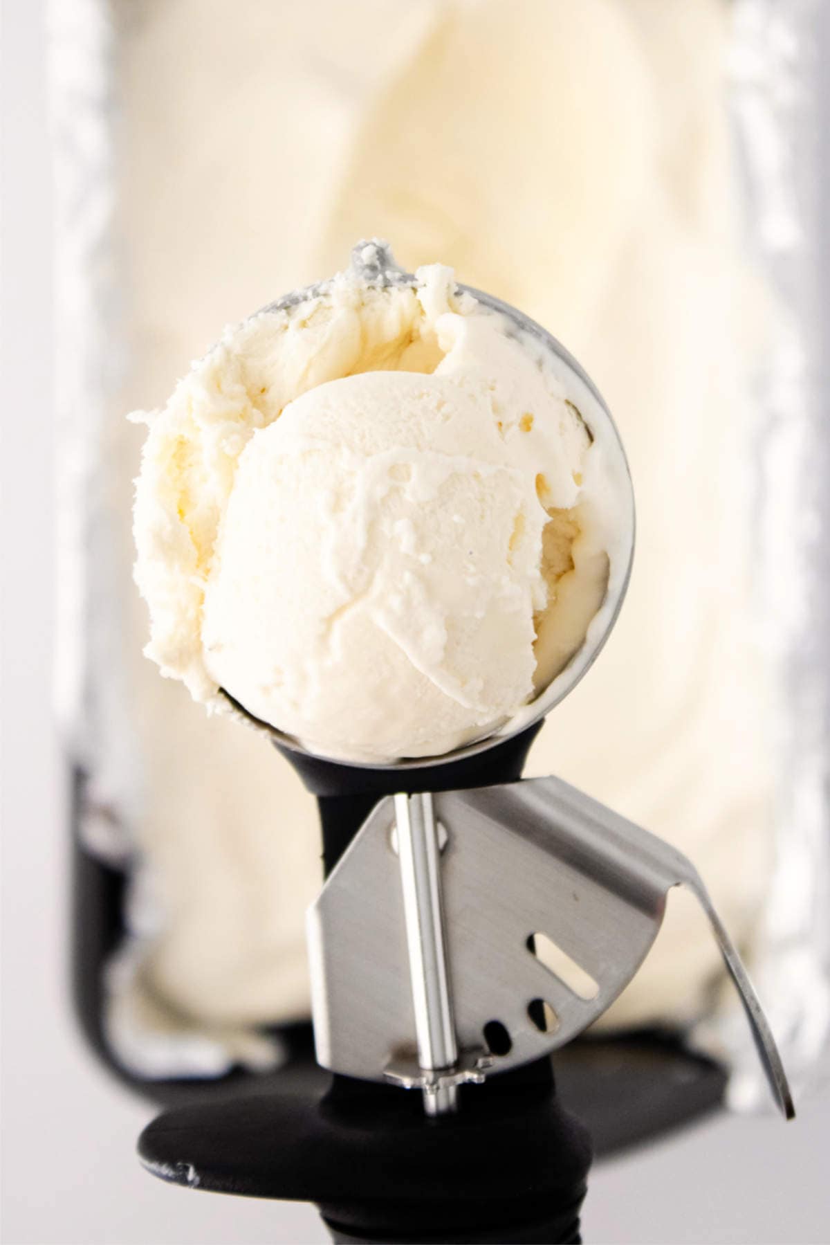 A metal ice cream scoop holds a rounded scoop of creamy vanilla ice cream, freshly made with an ice cream maker, with more vanilla ice cream visible in a container in the blurred background.