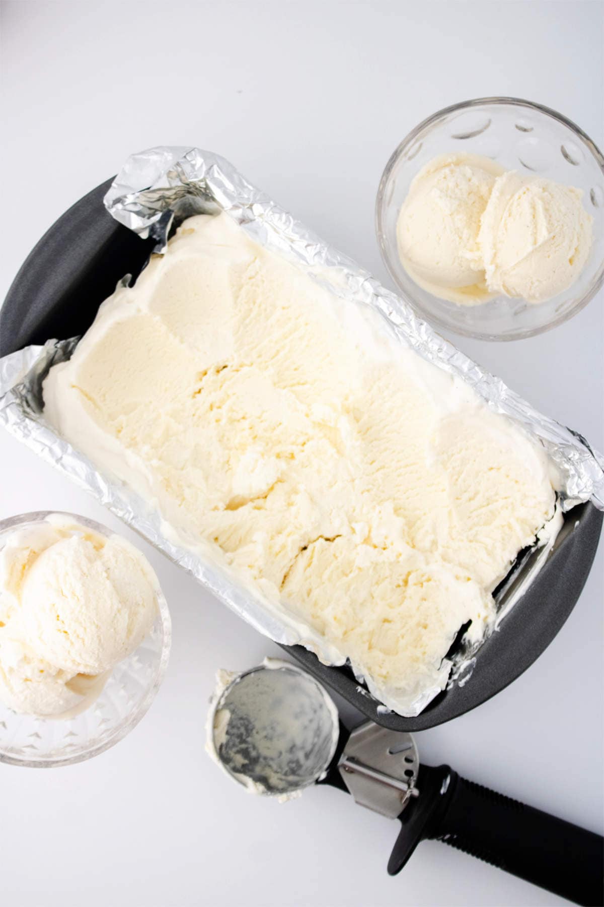 A loaf pan lined with foil filled with vanilla ice cream made from an ice cream maker, two glass bowls each with scoops of vanilla ice cream, and an ice cream scooper with some melted ice cream on it, all on a white background.