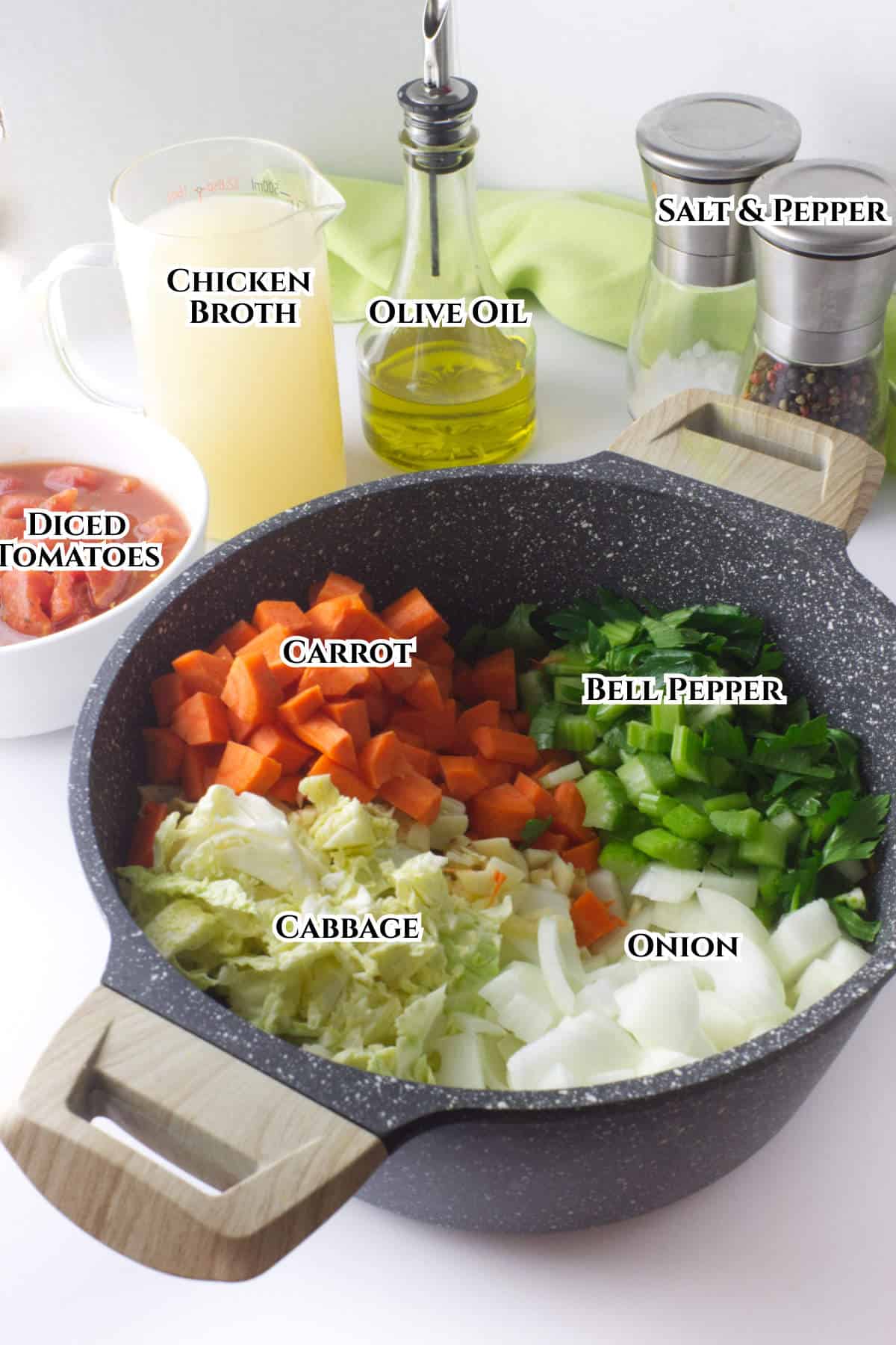 A pot with chopped carrots, cabbage, bell pepper, and onion, surrounded by a pitcher of chicken broth, a bottle of olive oil, a bowl of diced tomatoes, and salt and pepper shakers, each labeled.