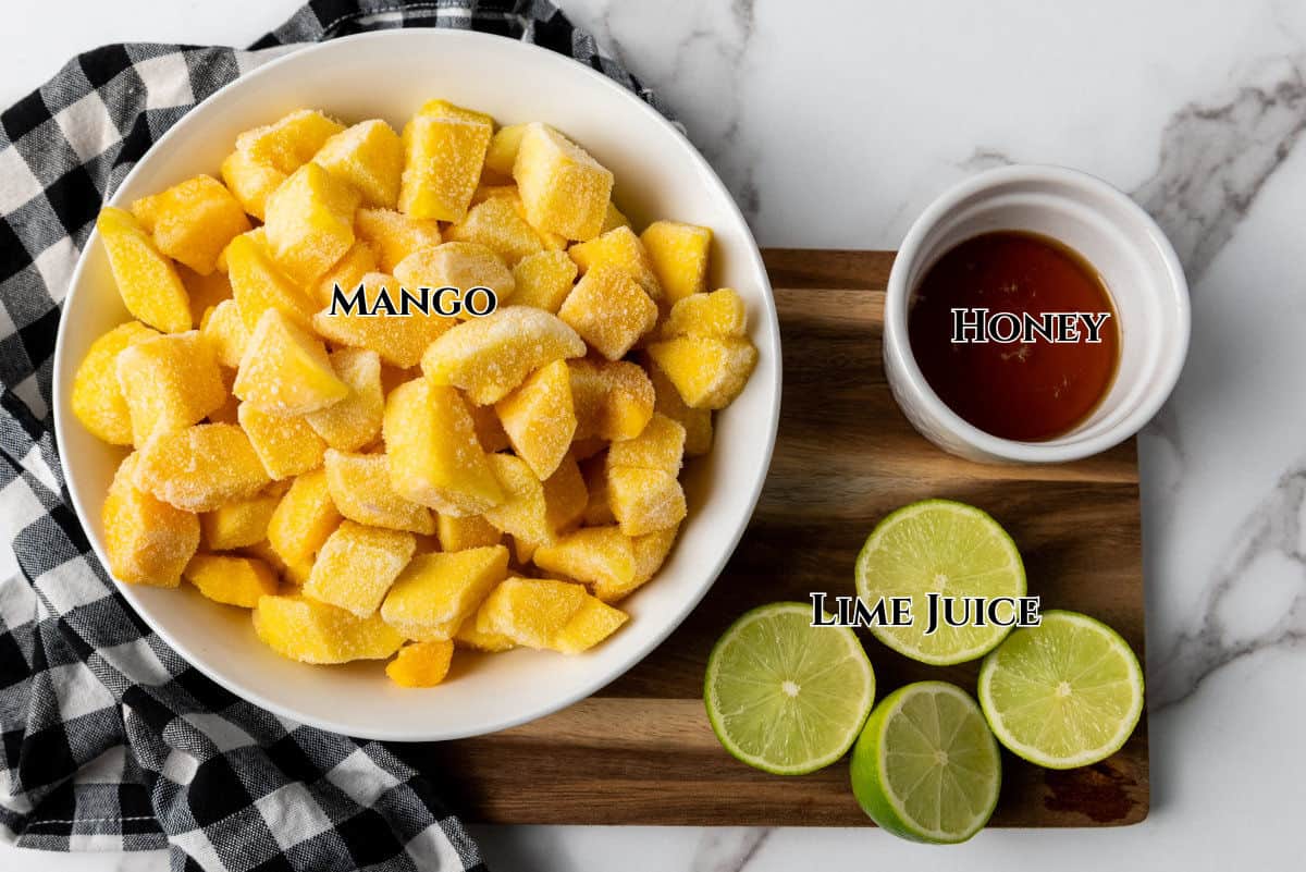 A bowl of frozen mango chunks, a small bowl of honey, and three lime halves on a wooden board with a black-and-white checkered cloth. Each ingredient is labeled: Mango, Honey, and Lime Juice.