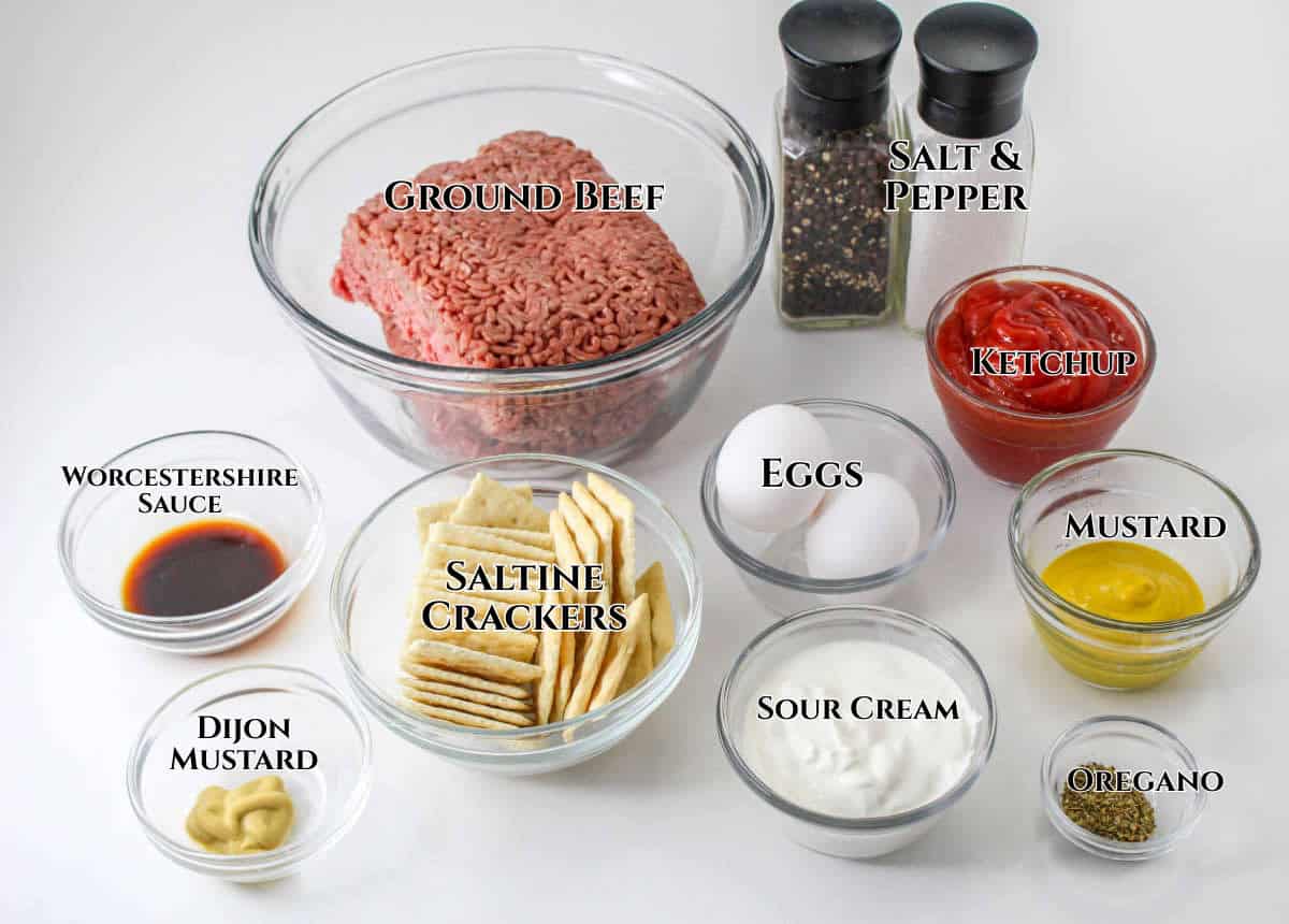 A variety of southern style meatloaf ingredients in bowls: ground beef, salt and pepper, ketchup, eggs, mustard, sour cream, oregano, Dijon mustard, Worcestershire sauce, and saltine crackers.