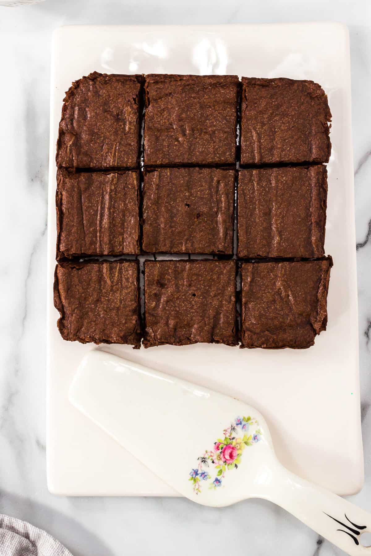 Nine one bowl brownies cut into squares are arranged in a 3x3 grid on a white rectangular tray. A white cake server with a floral design rests next to the brownies on the tray.