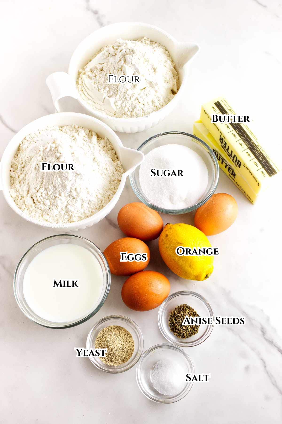 Two bowls of flour, a stick of butter, a bowl of sugar, three eggs, a glass of milk, an orange, bowls of yeast, anise seeds, and salt are arranged on a white surface, each labeled with its name.