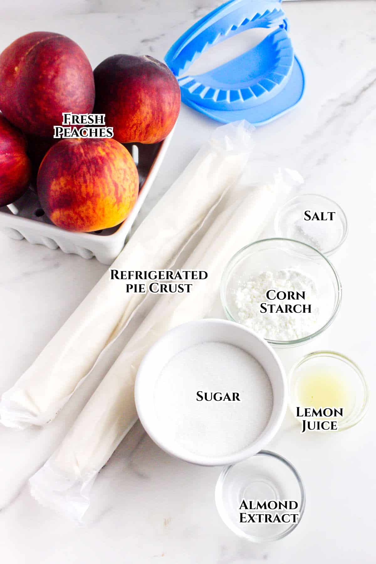 Ingredients for peach hand pies are arranged on a white surface: fresh peaches, refrigerated pie crust, sugar, almond extract, lemon juice, corn starch, salt, and a blue plastic hand pie mold.