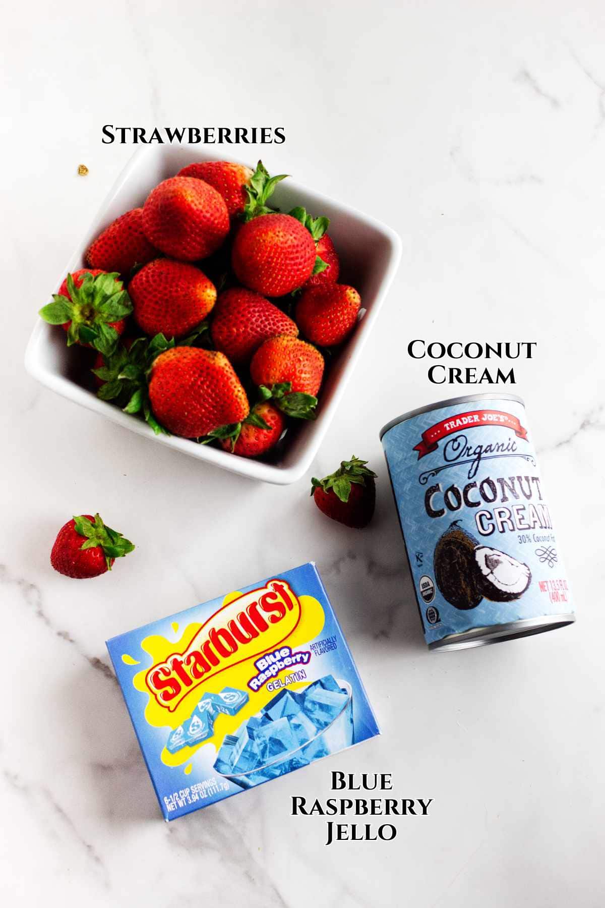 A bowl of fresh strawberries, a can of Trader Joe's organic coconut cream, and a box of Starburst blue raspberry jello on a white surface, each labeled with text.