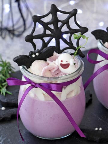 A glass jar of purple mousse topped with frosting, a smiling ghost-shaped candy, a chocolate bat, and a chocolate spiderweb. Tied with a purple ribbon, this is one of the cutest halloween treats for a festive celebration.