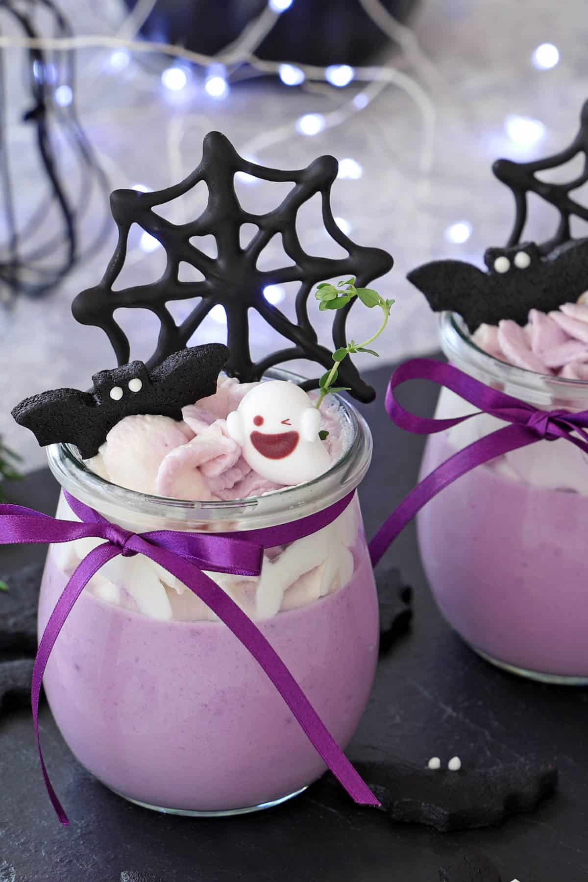 A jar of purple mousse is decorated with violet ribbon, black cookie bats, a spiderweb cookie, and a white marshmallow with a smiling face. Blurred fairy lights in the background complete these festive halloween treats.