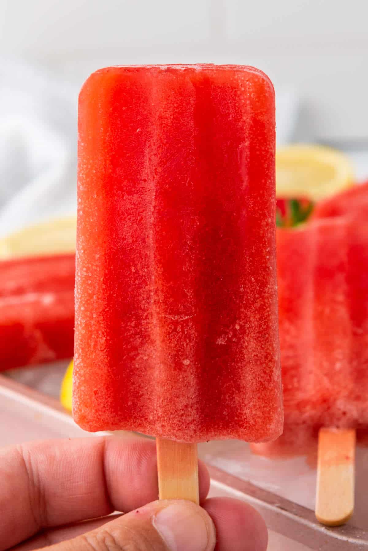 A close-up of a hand holding a bright red popsicle with a wooden stick, with more popsicles and slices of lemon and strawberry blurred in the background.