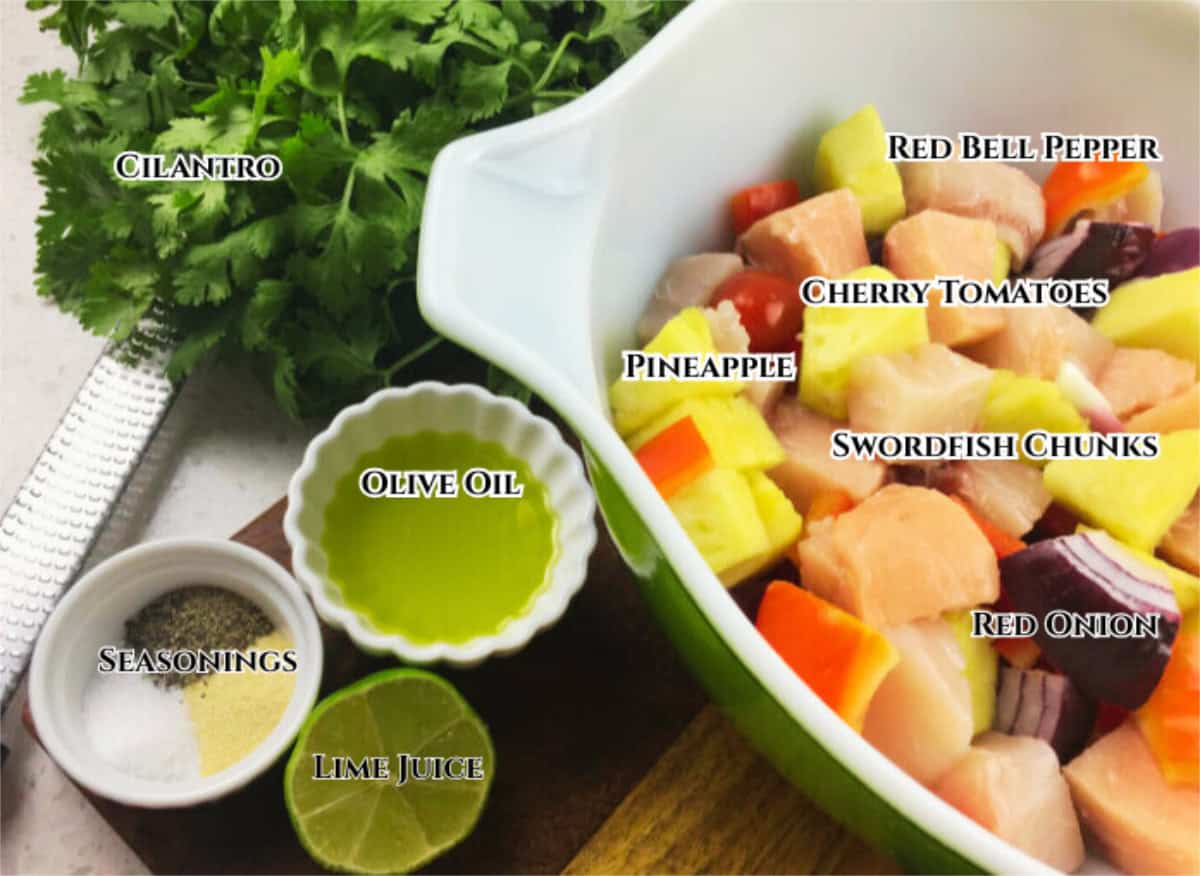 A bowl with swordfish kabobs ingredients like swordfish chunks, pineapple, red bell pepper, cherry tomatoes, and red onion sits next to cilantro, olive oil, lime juice, and seasonings on a wooden board with ingredient labels.