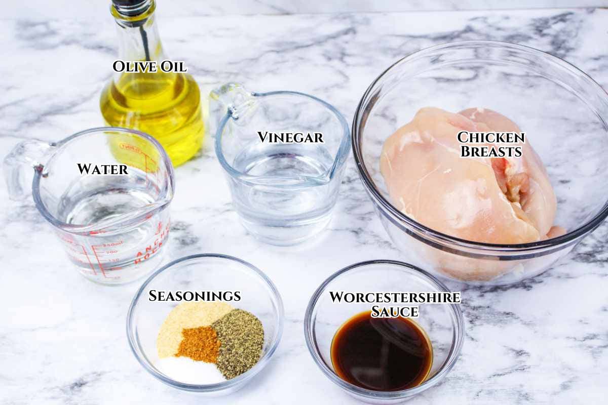 A marble countertop displays bowls and measuring cups containing olive oil, vinegar, raw chicken breasts, water, Worcestershire sauce, and assorted seasonings, each container labeled with its contents.
