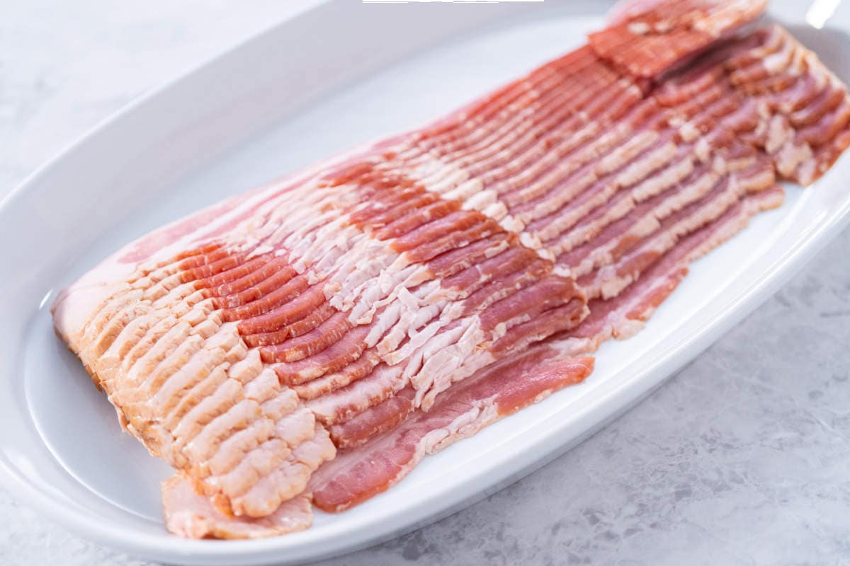 Several slices of raw bacon are neatly arranged on a large white oval plate, ready to be transformed into crispy air fryer bacon on a light-colored marble surface.