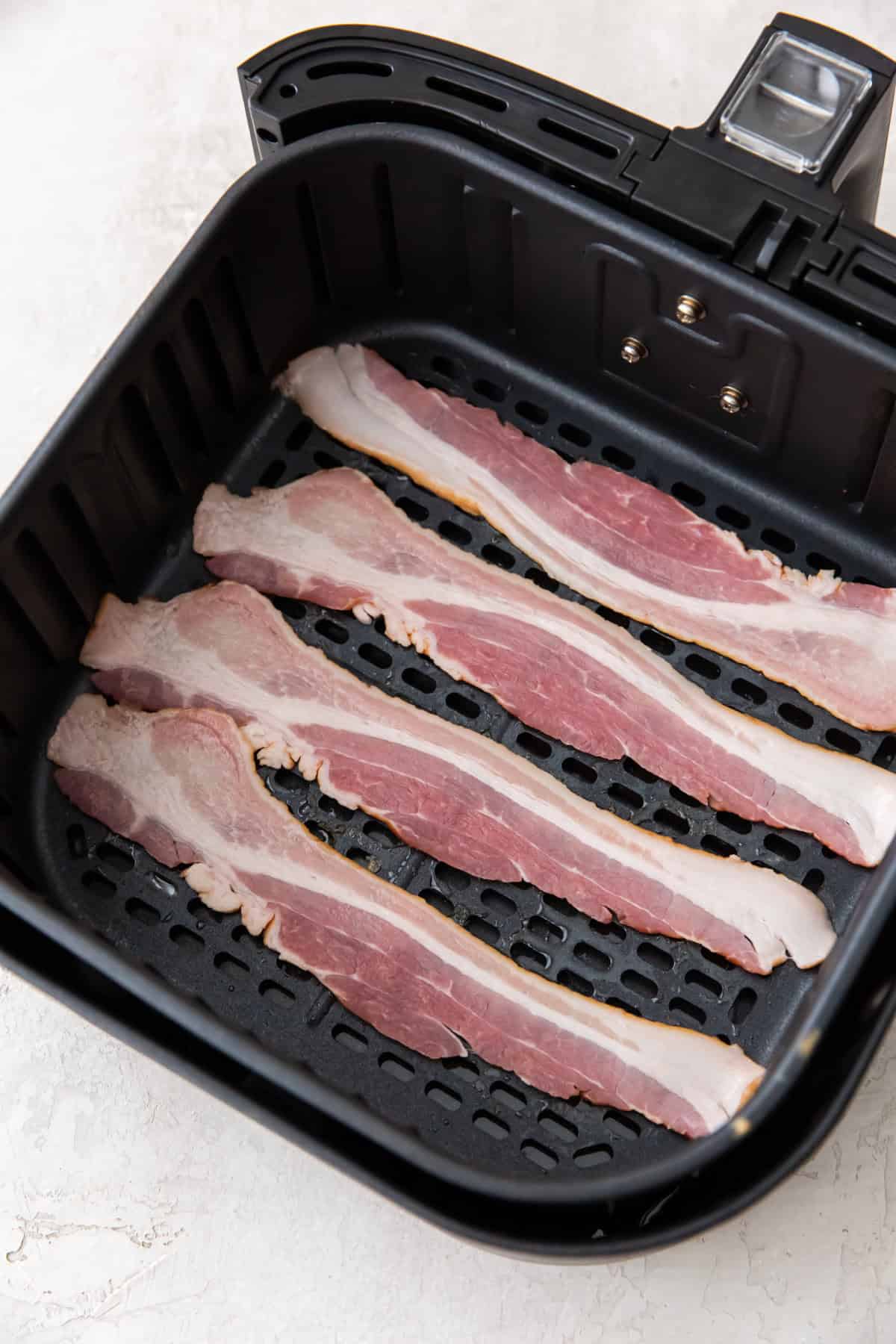 Four raw strips of bacon are placed in a single layer inside the basket of a black air fryer, ready to become crispy air fried bacon. The background is a light-colored countertop.