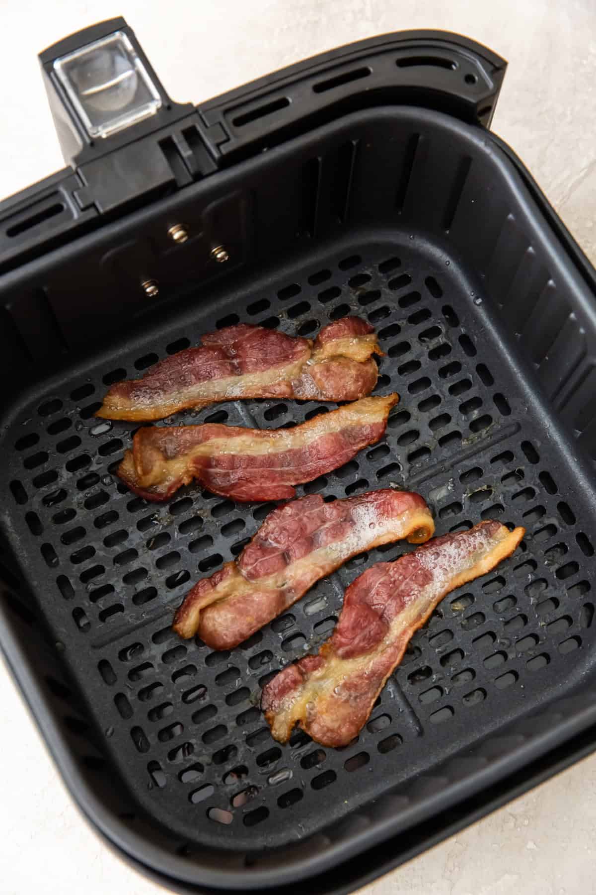 Four cooked strips of air fried bacon sit inside the basket of a black air fryer, resting on a perforated tray with a light-colored countertop in the background.