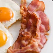 Two sunny-side-up eggs and several strips of crispy air fried bacon are arranged on a white plate, making a classic breakfast combination.