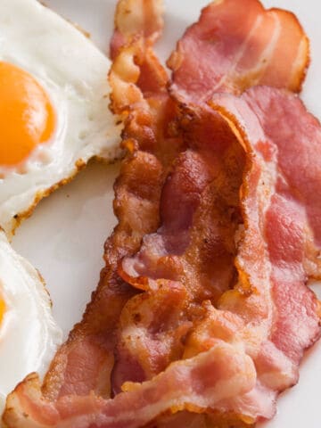 Two sunny-side-up eggs and several strips of crispy air fried bacon are arranged on a white plate, making a classic breakfast combination.