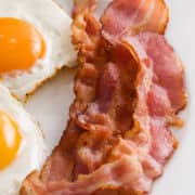 Two sunny-side-up eggs and three crispy strips of air fried bacon are served on a white plate.