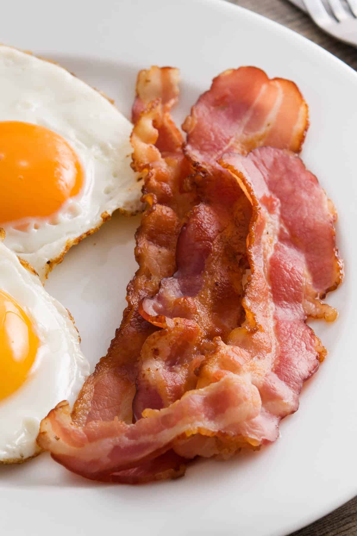 Two sunny-side-up eggs and three crispy strips of air fried bacon are served on a white plate.