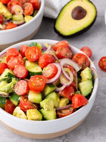 A bowl of Avocado Cucumber and Tomato Salad with cherry tomatoes, avocado, cucumber, and red onion slices rests on a gray surface. A halved avocado and another bowl of salad appear in the background.