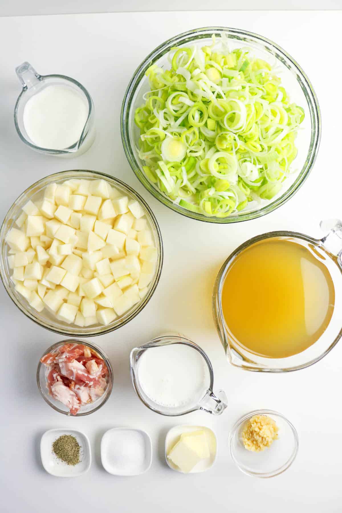 Top-down view of ingredients for bacon potato and leek soup in bowls and measuring cups: sliced leeks, diced potatoes, broth, bacon pieces, milk, cream, butter, minced garlic, salt, and pepper—arranged neatly on a white surface.