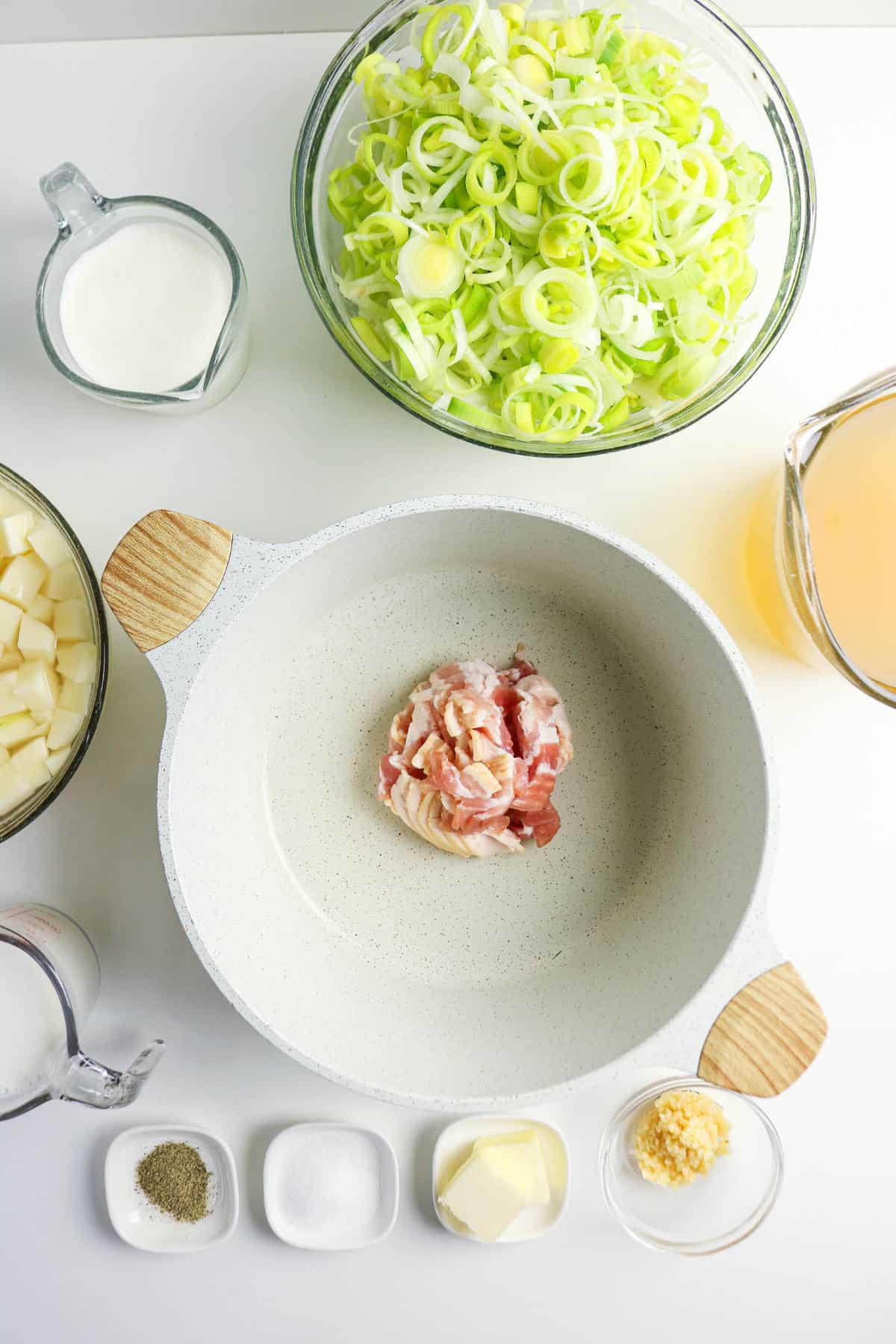 Top-down view of a white pot with chopped raw bacon, surrounded by bowls of sliced leeks and diced potatoes-ingredients for a comforting bacon potato and leek soup-plus a pitcher of broth, milk, cream, and small dishes with butter, garlic, salt, and pepper.