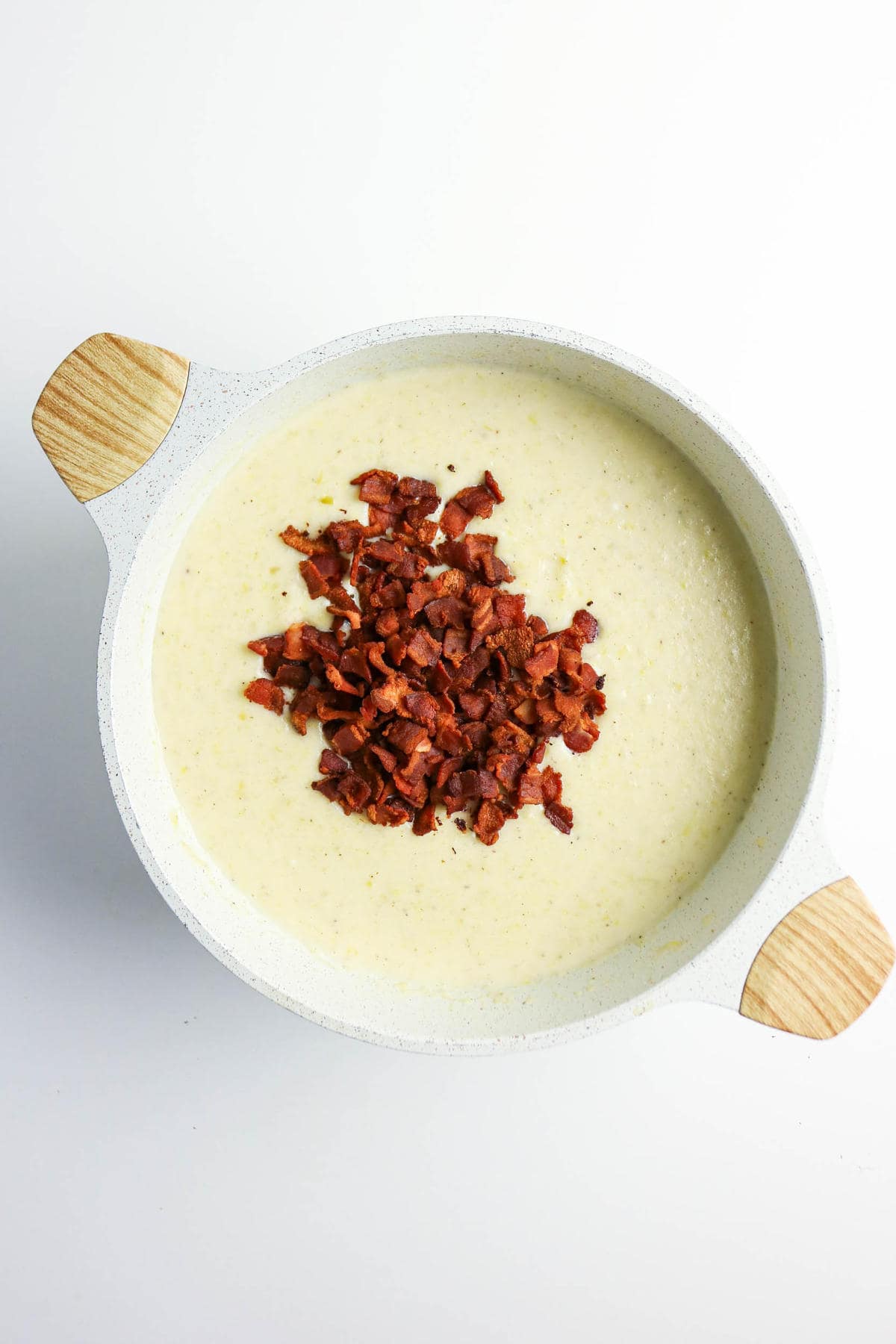 A white pot with wooden handles filled with creamy bacon, potato, and leek soup, topped with a pile of crispy chopped bacon pieces, set against a white background.