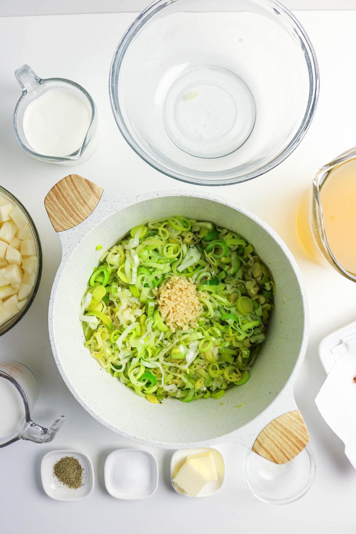 A white pot filled with sliced leeks and minced garlic sits on a white surface, ready for bacon potato and leek soup. Surrounding the pot are bowls of diced potatoes, milk, broth, butter, cream, and dishes of salt and pepper.