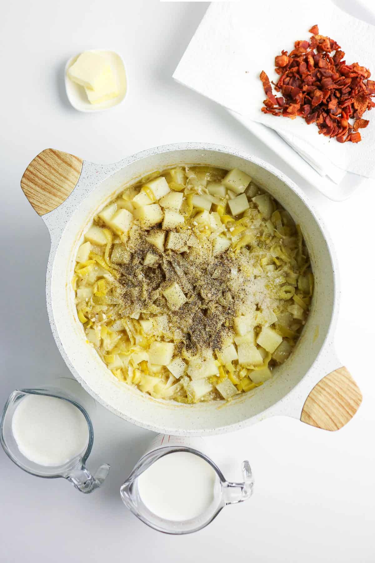 A pot filled with chopped potatoes, onions, and spices sits on a white surface-perfect beginnings for bacon potato and leek soup. Nearby are two measuring cups with milk, a pat of butter on a small plate, and crumbled bacon on a paper towel.