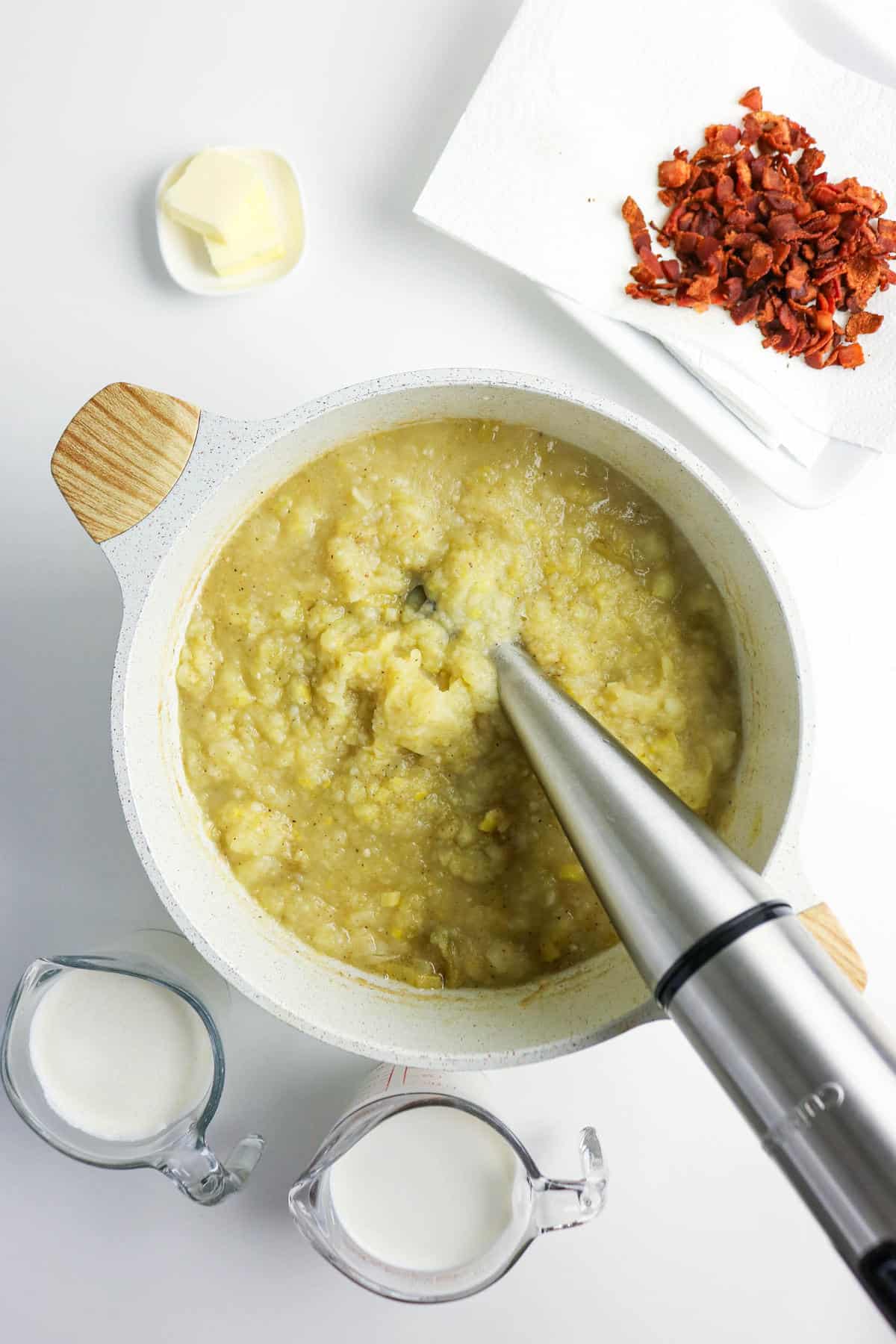A hand blender purees cooked potatoes in a pot for bacon potato and leek soup. Nearby are a small dish of butter, chopped cooked bacon on a paper towel, and two measuring cups filled with milk or cream.