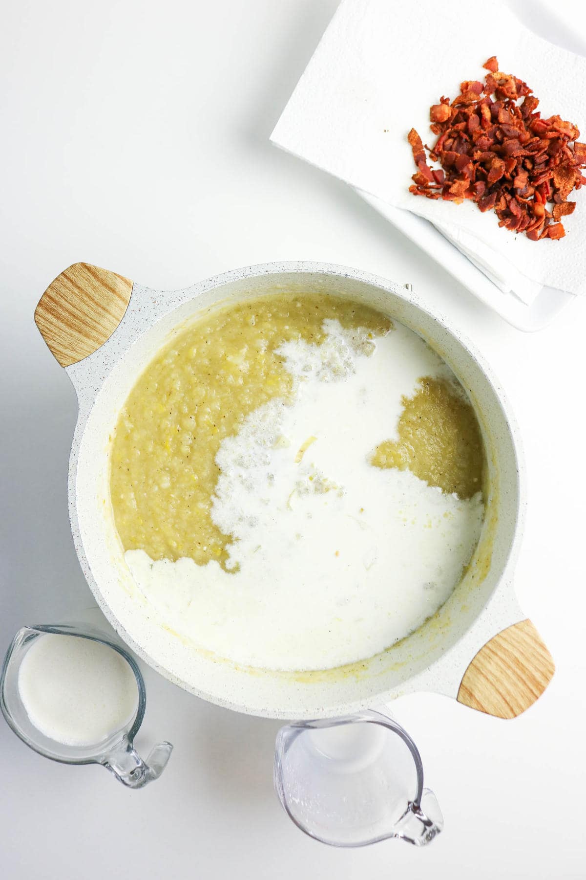 A pot of creamy bacon potato and leek soup with milk being added sits on a white surface. Nearby are two measuring cups-one with milk, one empty-and a paper towel topped with chopped cooked bacon.