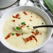 A bowl of creamy bacon potato and leek soup garnished with crispy bacon pieces and chopped green onions, with a spoon resting inside. A checkered cloth and part of a leek are visible in the background.