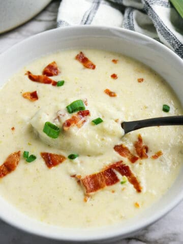 A bowl of creamy bacon potato and leek soup garnished with crispy bacon pieces and chopped green onions, with a spoon resting inside. A checkered cloth and part of a leek are visible in the background.
