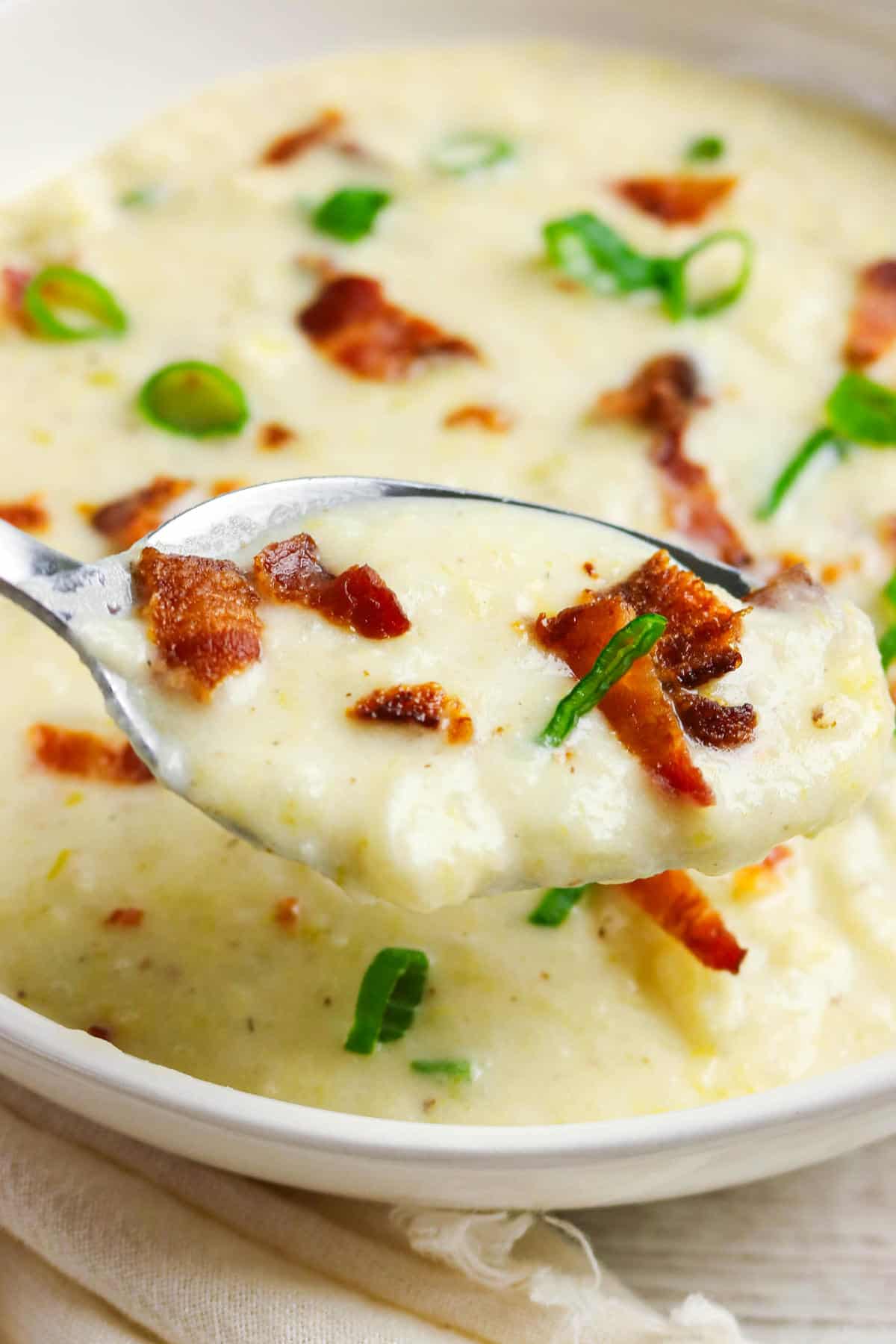A close-up of a spoonful of creamy bacon potato and leek soup, topped with crispy bacon bits and sliced green onions, held above a bowl filled with more of the savory soup.